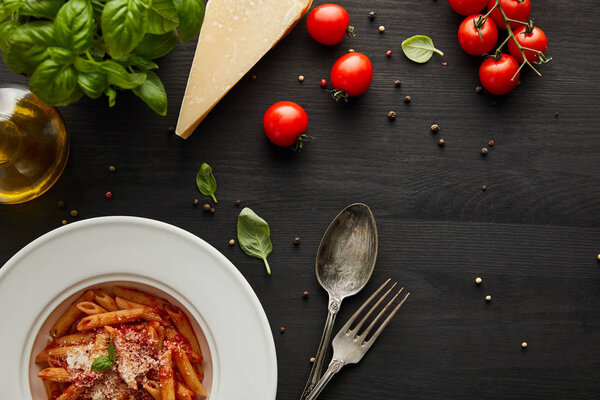 top view of tasty bolognese pasta with tomato sauce and Parmesan in white plate near ingredients and cutlery on black wooden background