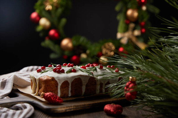 селективный фокус традиционного рождественского торта с клюквой возле рождественского венка с безделушками на деревянном столе, изолированном на черном
