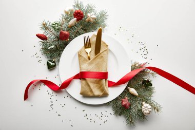 Peçeteli, çatal bıçaklı ve kurdeleli beyaz tabağın üst görüntüsü beyaz arka planda ziyafet dolu Noel ağacı dalları ve konfetileri.