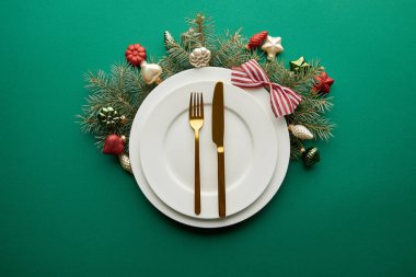 Beyaz tabakların üst görüntüsü, yeşil arka planda mücevherleri olan şenlikli Noel ağacı dalının yanındaki çatal bıçak takımı.
