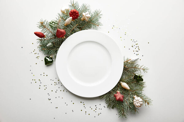 верхний вид праздничного накрытия Рождественского стола на белом фоне с украшенной сосновой ветвью, тарелкой и конфетти
