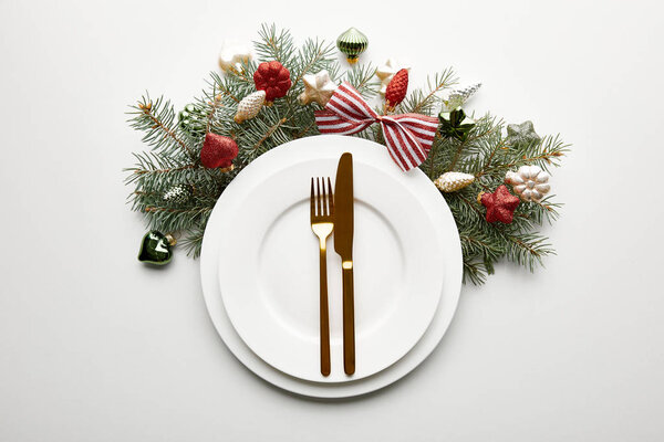 верхний вид на белые тарелки со столовыми приборами рядом с праздничной веткой елки с безделушками на белом фоне
