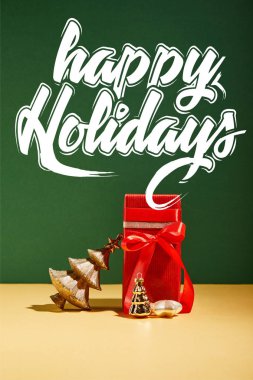 Kırmızı hediye kutusu ve dekoratif Noel ağacı. Yeşil arka planda altın mücevherler, beyaz mutlu bayramlar yazıları.