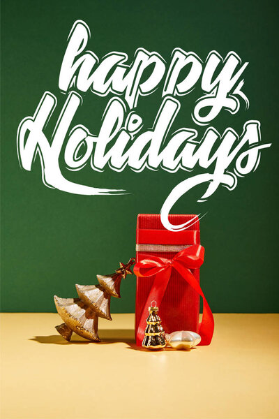 red gift box and decorative Christmas tree with golden baubles on green background with white happy holidays lettering