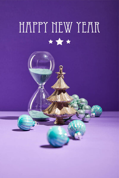 selective focus of decorative Christmas near blue baubles and hourglass on purple background with happy new year lettering