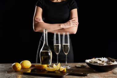 cropped view of woman standing with crossed arms near sparkling wine in bottle and oysters in bowl with ice isolated on black  clipart
