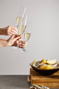 cropped view of women clinking champagne glasses with sparkling wine near bowl with oysters and fresh lemons isolated on grey  clipart