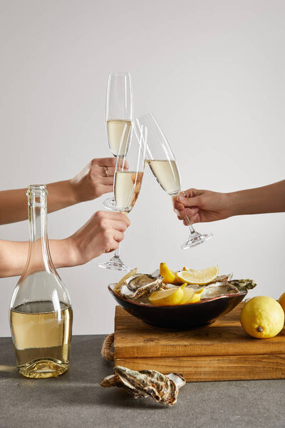cropped view of women toasting champagne glasses with sparkling wine near bowl with oysters and lemons isolated on grey 
