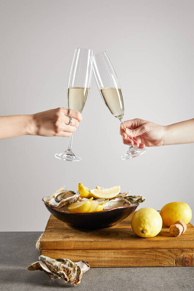 cropped view of women toasting champagne glasses with sparkling wine near bowl with oysters and fresh lemons isolated on grey 