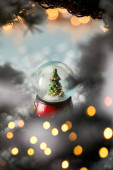 malá sněhová koule s vánočním stromečkem stojící na modré se smrkovými větvemi, sněhem a rozmazanými světly 
