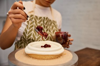 Cropped view of confectioner adding jam on sponge cake with cream  clipart