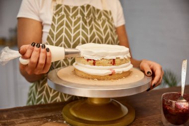Cropped view of confectioner putting cream from pastry bag on sponge cake clipart