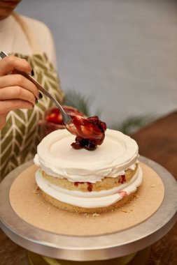 Cropped view of confectioner with spoon putting berry jam on cake with cream clipart