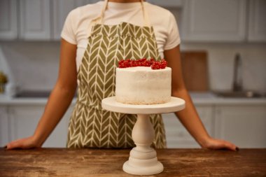 Cropped view of confectioner standing beside tasty cake with redcurrant on table clipart