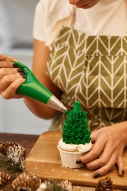 Cropped view of confectioner with pastry bag decorating Christmas tree cupcake on table with spruce cones  clipart