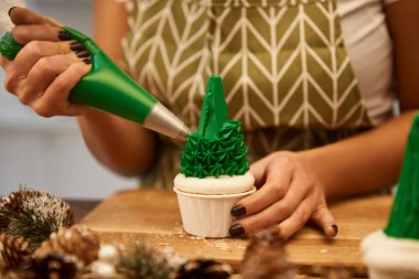 Cropped view of confectioner decorating Christmas tree cupcake with green cream on table with spruce cones  clipart