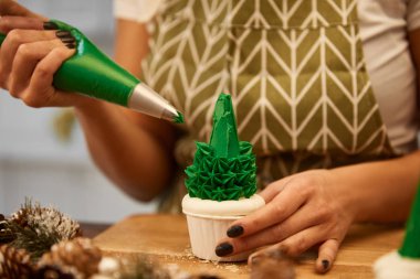 Cropped view of confectioner adding green cream on Christmas tree cupcake beside spruce cones on table clipart