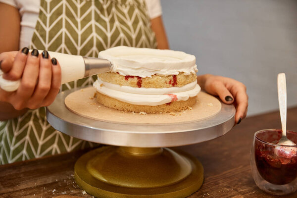 Cropped view of confectioner adding cream on cake layers with berry jam 