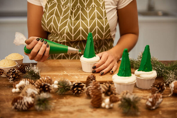 Cropped view of confectioner decorating cupcake with pastry bag beside pine cones on table