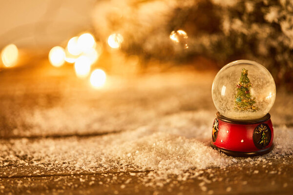 маленький снежок с рождественской ёлкой, стоящей на деревянном столе в снегу с еловыми ветвями и размытыми огнями ночью
 