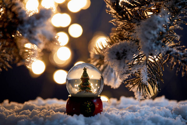 декоративная елка в снежке, стоящая в снегу с еловыми ветвями и размытыми огнями ночью
