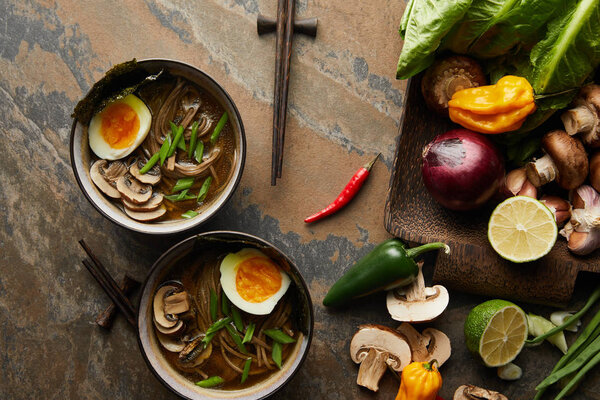 top view of traditional spicy ramen in bowls near chopsticks and vegetables on stone surface