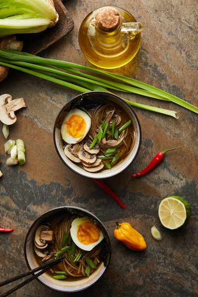 top view of traditional spicy ramen in bowls with chopsticks near vegetables  and oil on stone surface