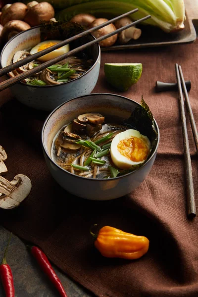 traditional spicy ramen in bowls with chopsticks and vegetables on brown napkin on stone surface