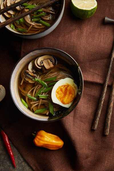 top view of traditional spicy ramen in bowls with chopsticks and vegetables on brown napkin on stone surface