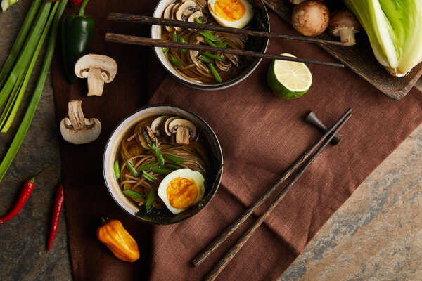 top view of traditional spicy ramen in bowls with chopsticks and vegetables on brown napkin on stone surface