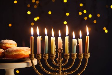 delicious doughnuts on stand near glowing candles in menorah on black background with bokeh lights on Hanukkah clipart