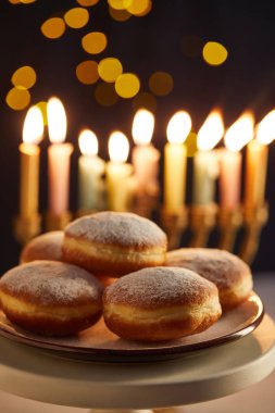 selective focus of delicious doughnuts on stand near glowing candles on black background with bokeh lights on Hanukkah clipart
