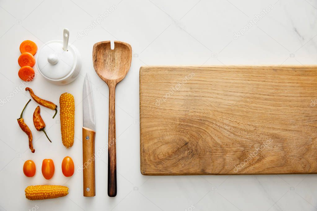 Top view of cutting board with kitchenware and vegetables on marble background