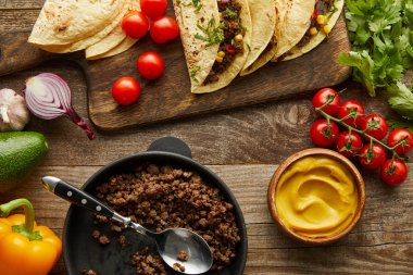 Top view of delicious tacos with minced meat on cutting board and ingredients on wooden background clipart