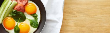 top view of fried eggs with spinach leaves, cucumber and sausage in frying pan on napkin on wooden table, panoramic shot clipart