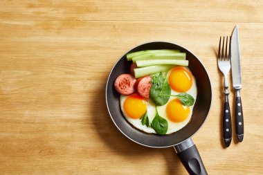 Kızarmış yumurta, ıspanak yaprakları, salatalık ve sosis, tavada çatal ve bıçağın yanında ahşap masa.