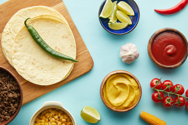 Top view of tortillas on cutting board with taco ingredients on blue background