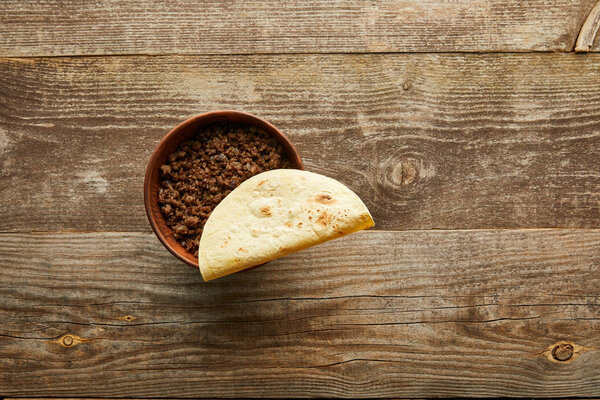 Top view of tortilla on bowl with minced meat on wooden background