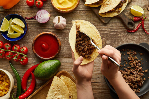 Cropped view of man adding minced meat in taco with raw ingredients on wooden surface