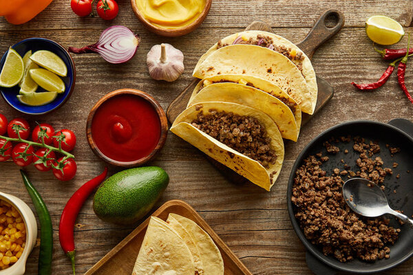 Top view of minced meat, tacos and raw ingredients on wooden background