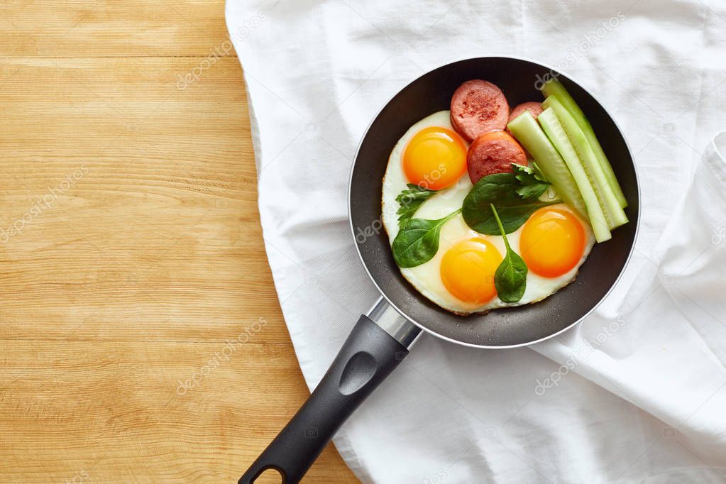 top view of fried eggs with spinach leaves, cucumber and sausage in frying pan on napkin on wooden table