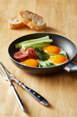 Kızartma tavasında yumurta, ıspanak, salatalık ve sosis, ahşap masada çatal bıçak ve ekmek ile seçici bir odak noktası.