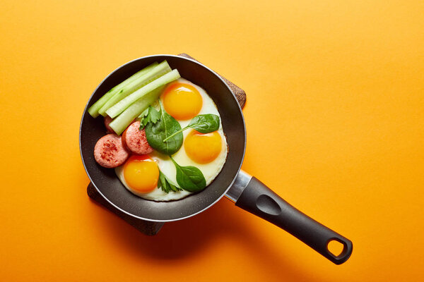 верхний вид жареных яиц со шпинатными листьями, огурцом и колбасой в сковороде на оранжевом фоне
