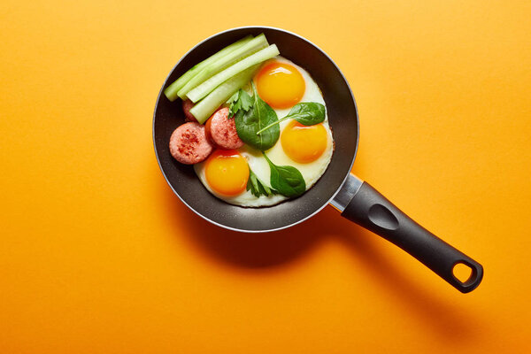 top view of fried eggs with spinach leaves, cucumber and sausage in frying pan on orange background