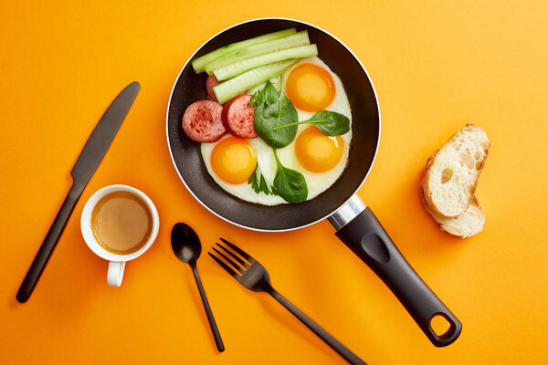 top view of fried eggs with spinach leaves, cucumber and sausage in frying pan near coffee, cutlery and bread on orange background