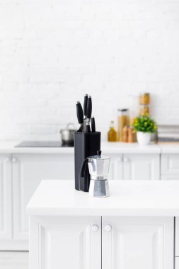 Modern beyaz mutfak masasında kahve demliği ve bıçaklar var.