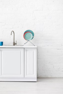 Modern beyaz mutfak. Tuğla duvarın yanında lavabo ve sofra takımı var.