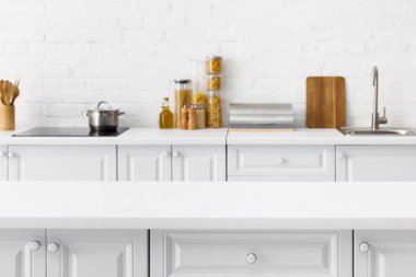 Boş masa üstünün seçici odağı ve arka plandaki tuğla duvarın yanında mutfak gereçleri ve yiyecekleri olan minimalist modern beyaz mutfak.