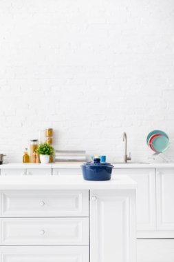 Mavi tencerenin seçici odak noktası ve arka planda mutfak eşyaları ve tuğla duvarın yanında yiyecek olan minimalist modern beyaz mutfak.