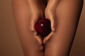 oříznutý pohled na ženu v nylonových punčocháčích držící zralé červené jablko izolované na hnědé